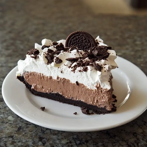 A slice of chocolate cream pie topped with whipped cream and crushed Oreo cookies on a plate.