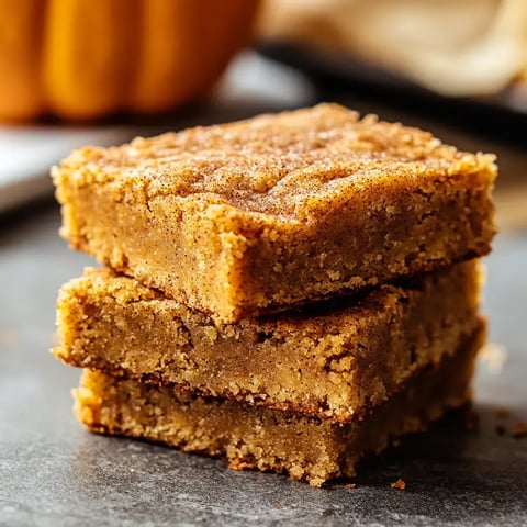 A stack of three golden-brown pumpkin bars sits on a textured surface, with a blurred pumpkin in the background.